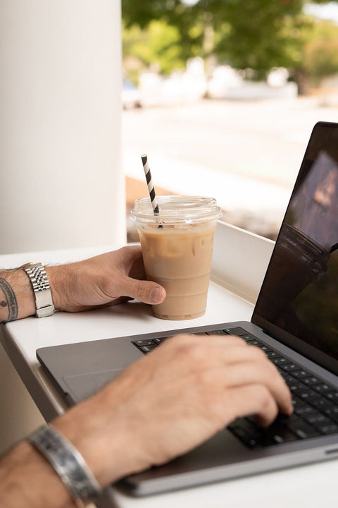 Person using a laptop with an iced coffee on a table outdoors.
