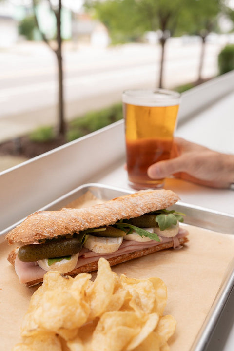 Sandwich with pickles, a glass of beer, and potato chips on a tray outdoors.