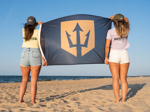 Two people holding a flag with a bold mariner trident logo on a beach