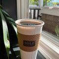 Coffee cup with a brown sleeve labeled 'Bold' on a windowsill with a view of greenery outside.