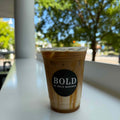 Iced coffee in a cup with 'BOLD' branding on a white surface with a blurred outdoor background