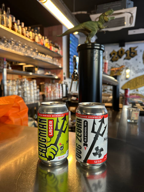 Two cans of Frogman Lager and Dog Zebra on a bar counter with a bar setting in the background.