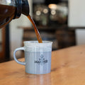 Coffee being poured into a mug with 'Bold Mariner' branding on a wooden table.