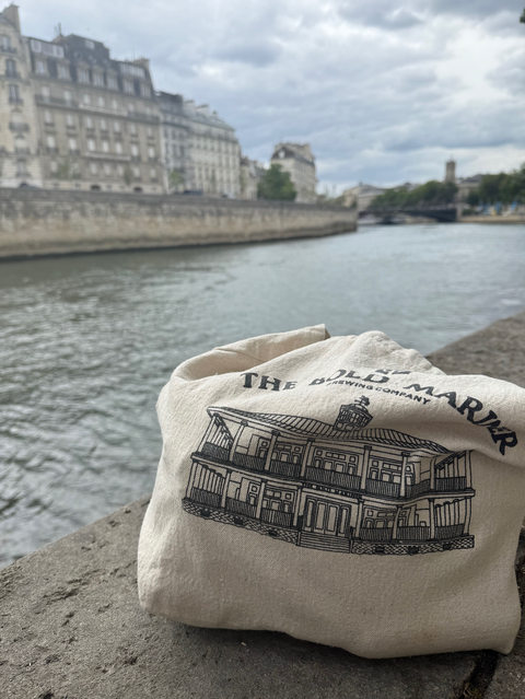 Bold Mariner beige tote bag with black logo and text by a river with buildings in the background in London