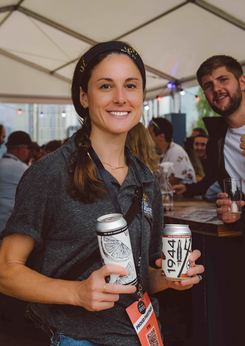 Woman holding two cans at an outdoor event with a tent in the background