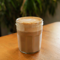 Glass jar with a hot cortado on a wooden surface with a blurred plant background