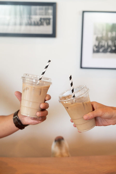 Two hands holding iced coffee drinks with straws against a blurred indoor background