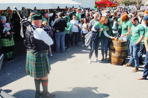 Person in a kilt playing bagpipes at a lively outdoor event with people in green shirts.