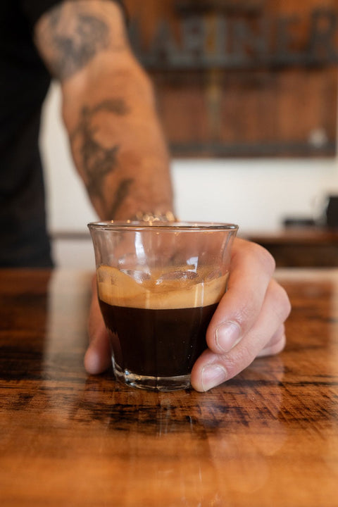 Hand holding a glass of espresso on a wooden surface with a blurred background.