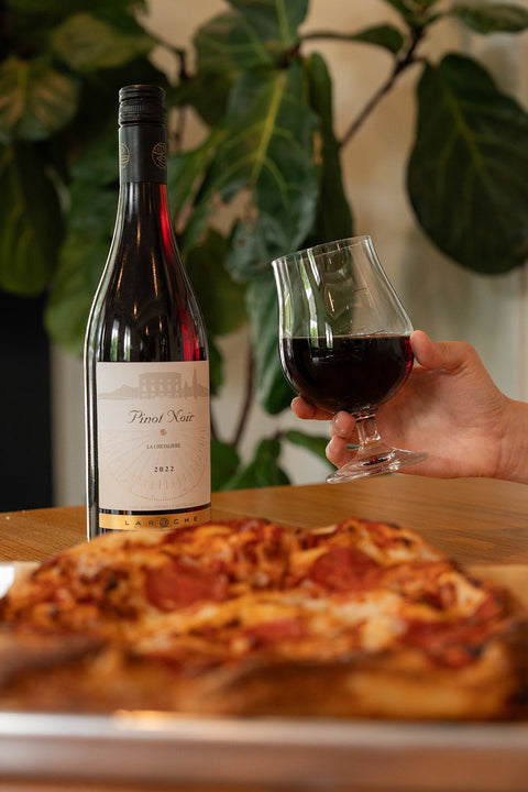 Bottle of wine, glass of wine, and pizza on a wooden table with a blurred plant background.