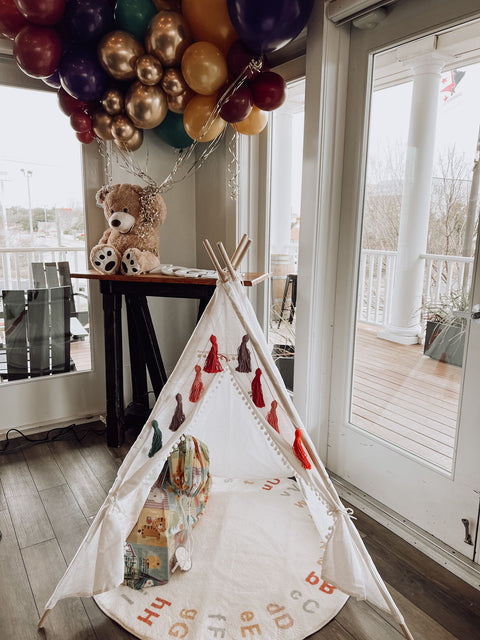 Children's teepee with colorful balloons and teddy bear in a room with large windows.