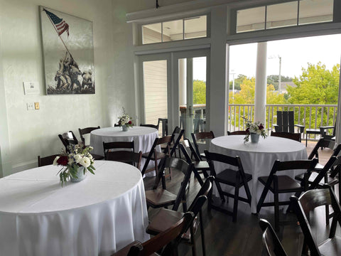 Dining room with round tables, chairs, and floral arrangements near a large window.