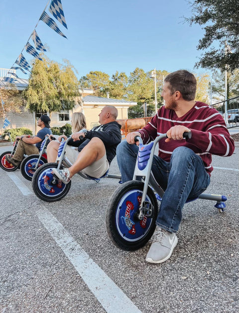 People riding small bikes in a parking lot with trees and buildings in the background.
