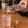Person pouring pink Luminia sparkling pink lemonade into a glass on a wooden table.