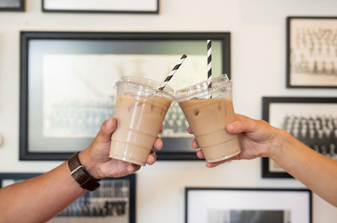 Two hands clinking iced coffee drinks with straws against a wall with framed pictures.