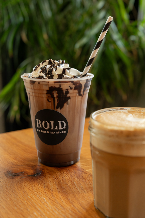 Two coffee drinks on a wooden surface with a blurred green plant background