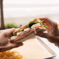 Two hands splitting a sandwich with chips on a tray.