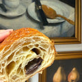 Close-up of a croissant with chocolate inside with a painting of pears in the background